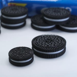 May include: A collection of round sandwich cookies featuring dark black textured cocoa wafers and a thin white cream filling. The cookies are arranged in stacks and individually, with a blue and white plastic packaging wrapper visible in the background.