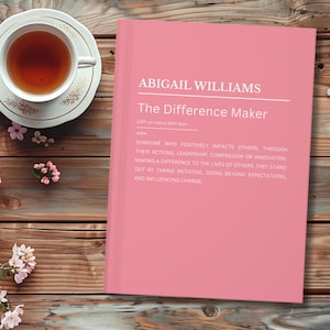 Puede incluir: Un libro rosa titulado "The Difference Maker" de Abigail Williams, con texto que describe el término. Una taza de té en un platillo floral y flores rosas sobre una superficie de madera.