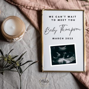 Könnte beinhalten: Eine goldgerahmte Ankündigungskarte mit weißem Hintergrund. Der Text lautet "We can't wait to meet you Baby Thompson March 2025". Im unteren rechten Eck befindet sich ein Schwarzweiß-Ultraschallbild.