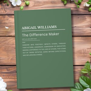 Puede incluir: Un libro verde titulado "The Difference Maker" de Abigail Williams, con una definición del término. El libro está sobre una superficie de madera, rodeado de flores blancas y hojas verdes.