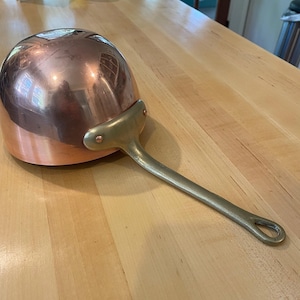 May include: A copper saucepan with a long, brass handle. The pan has a rounded body and a shiny, reflective surface. The handle is attached with rivets and has a loop at the end for hanging. Kitchenware.