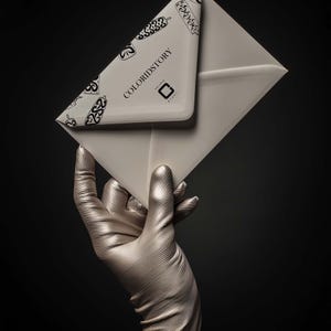 Op de afbeelding: Een metallic zilveren hand met een fijn gestreepte textuur houdt een crèmekleurige envelop vast. De envelopflap is voorzien van een herhalend zwart grafisch patroon en de tekst COLORIDSTORY met een klein vierkant logo eronder.