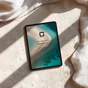 Puede incluir: Una maqueta de iPad con marco dorado muestra una escena de playa con agua turquesa y una orilla arenosa. La pantalla muestra el texto "IPAD MOCKUP PSD SMART LAYER" y un logotipo. La tableta descansa sobre una superficie clara y texturizada con sombras y tela blanca.