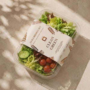 May include: A clear plastic container filled with a salad of mixed greens, sliced cucumbers, and cherry tomatoes. A paper label wraps around the center, printed with the text COLORIDSTORY CLEAN GREEN, listing ingredients like quinoa and chickpeas.