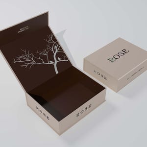 以下が含まれることがあります： 内側に白い木と鳥のデザインが施された茶色の箱。箱は開いており、蓋は後ろに折り畳まれています。箱の外側と内側に「ROSE」とラベルが貼られています。箱は白い表面の上に置かれています。