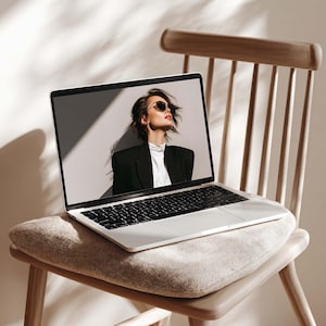 May include: A silver laptop with a black keyboard sits on a wooden chair with a beige cushion. The laptop screen displays a woman wearing sunglasses and a black blazer. The chair has a light wood finish.