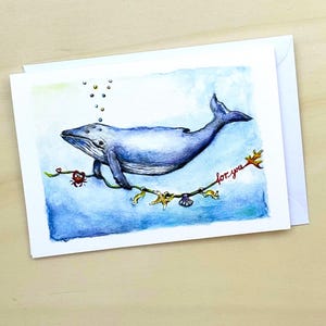 Könnte beinhalten: Ein Aquarellbild eines blauen Wals, der mit einer Schnur aus Muscheln und Seesternen schwimmt. Der Wal hält sich an einem Ast fest, an dem ein roter Krebs sitzt. Der Text "for you" ist in Rot auf dem Ast geschrieben.