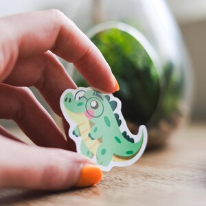 Peut inclure: Un autocollant découpé représentant un alligator de dessin animé portant des lunettes. L'alligator est principalement vert avec des reflets jaunes, une joue rose et des détails noirs. L'autocollant est tenu dans une main avec du vernis à ongles orange.