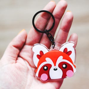 Może przedstawiać: Brelok z przezroczystego akrylu przedstawiający stylizowaną twarz pandy małej w żywych kolorach pomarańczowym, białym i ciemnoczerwonym. Postać ma duże oczy, różowe okrągłe policzki i spiczaste uszy. Przymocowany do czarnego metalowego kółka.