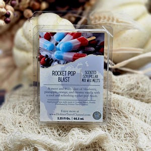 May include: A clear plastic clamshell container of Rocket Pop Blast scented soy and pillar mix wax melts. The label features red, white, and blue popsicle imagery, text describing the scent, and the website www.HolmesTravelChannel.com.