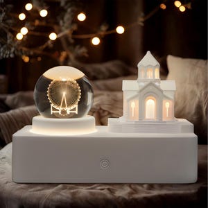 May include: A white decorative light with two illuminated features: a glass sphere with a Ferris wheel design and a church-shaped structure. The light sits on a rectangular base with a circular power button.