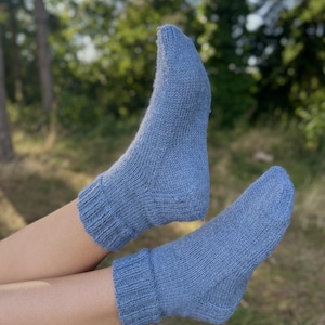 May include: A pair of light blue knitted socks. The socks have ribbed cuffs at the ankles and are made from a soft, textured yarn. The background is blurred, with green trees and sunlight.