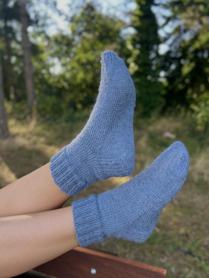 Pu&ograve; includere: Un paio di calzini lavorati a maglia blu chiaro. I calzini hanno polsini a coste alle caviglie e sembrano essere fatti di un filato morbido e strutturato. Lo sfondo &egrave; sfocato, con alberi verdi e luce solare.