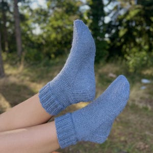 Pu&ograve; includere: Un paio di calzini lavorati a maglia blu chiaro. I calzini sono realizzati in un materiale morbido e strutturato e hanno polsini a coste. I calzini sono indossati sui piedi di una persona, con uno sfondo sfocato di alberi e verde.