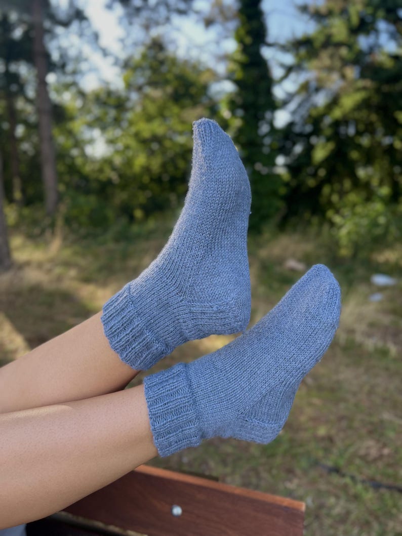 Pu&ograve; includere: Un paio di calzini lavorati a maglia blu chiaro. I calzini sono realizzati in un materiale morbido e strutturato e hanno polsini a coste. I calzini sono indossati sui piedi di una persona, con uno sfondo sfocato di alberi e verde.