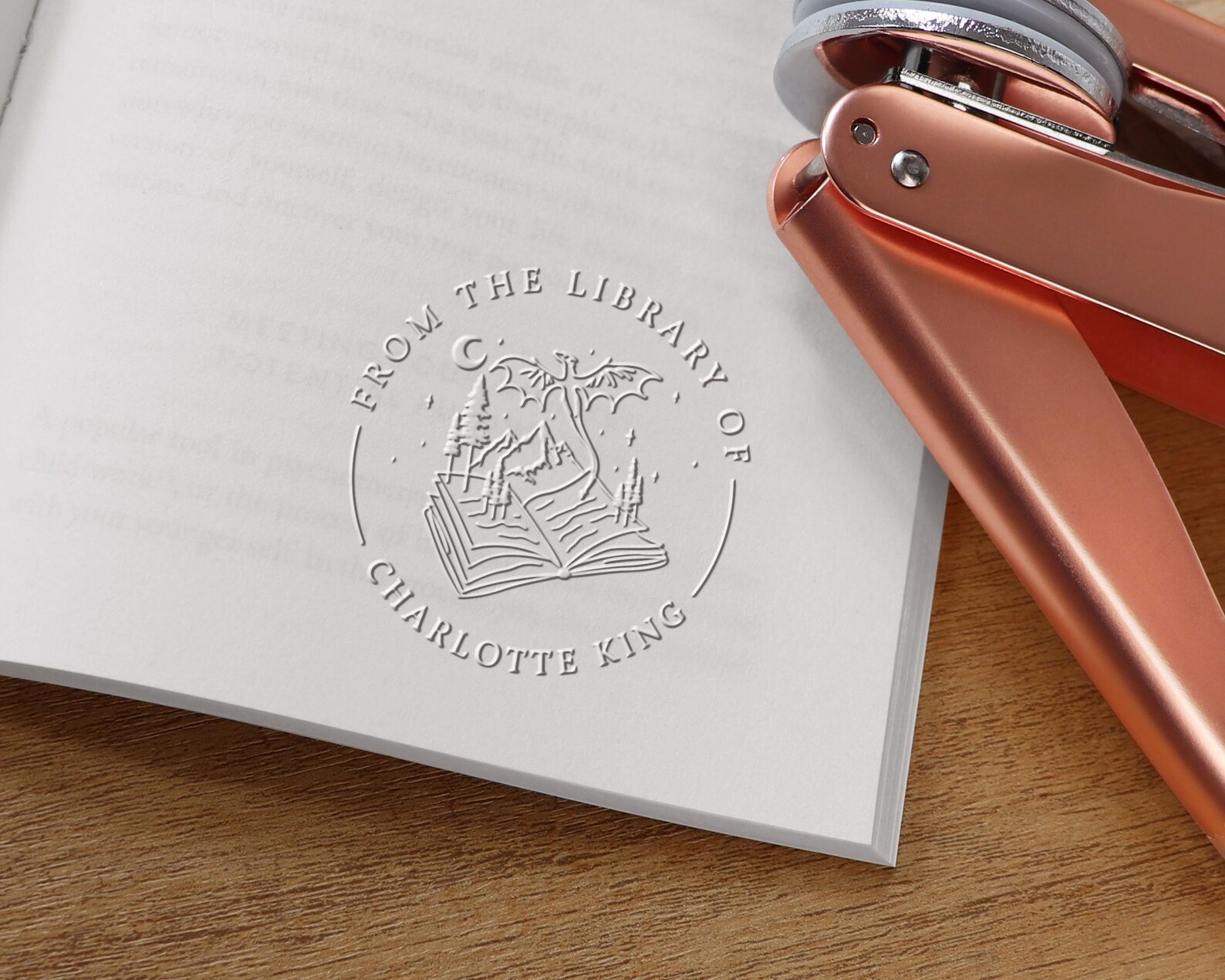 A metal embosser sits on the pages of a book embossed with "From the Library of Charlotte King" with an open book in the middle and a dragon emerging from the book on a background of trees, mountains, and the moon.