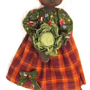 May include: A handmade rag doll with brown skin, black hair, and a green and red plaid dress. She is holding a green cabbage in her hands and is wearing a red and orange plaid headscarf.