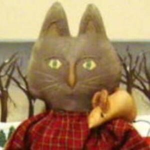 May include: A handcrafted, folk art cat doll with a whimsical design. The cat has a gray face, large green eyes, and a long nose. It wears a red plaid shirt and has a small, tan mouse companion. The background features a winter scene with bare trees.