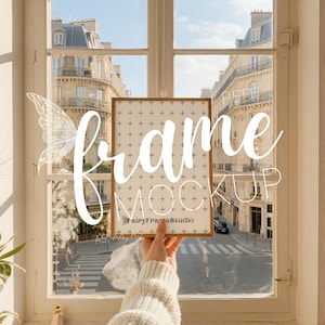 Peut inclure: Maquette de cadre tenue devant une fenêtre avec vue sur une rue européenne. Le cadre a une bordure en bois clair et un intérieur crème avec un motif quadrillé. Les mots "frame mockup" sont écrits en blanc.