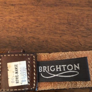 Puede incluir: Primer plano de un cintur&oacute;n de cuero marr&oacute;n con un borde cosido. El cintur&oacute;n tiene una etiqueta que dice "Brighton" en letras blancas sobre un fondo negro. Tambi&eacute;n es visible una etiqueta de precio.