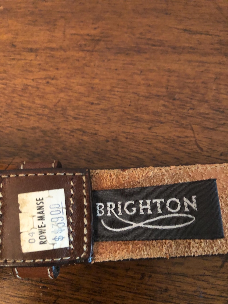 Puede incluir: Primer plano de un cintur&oacute;n de cuero marr&oacute;n con un borde cosido. El cintur&oacute;n tiene una etiqueta que dice "Brighton" en letras blancas sobre un fondo negro. Tambi&eacute;n es visible una etiqueta de precio.