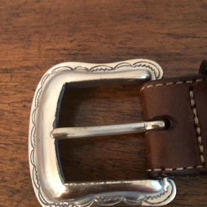 Puede incluir: Primer plano de una hebilla de cintur&oacute;n plateada con un borde festoneado decorativo. La hebilla est&aacute; sujeta a un cintur&oacute;n de cuero marr&oacute;n con costuras blancas. El dise&ntilde;o de la hebilla es de estilo western cl&aacute;sico.