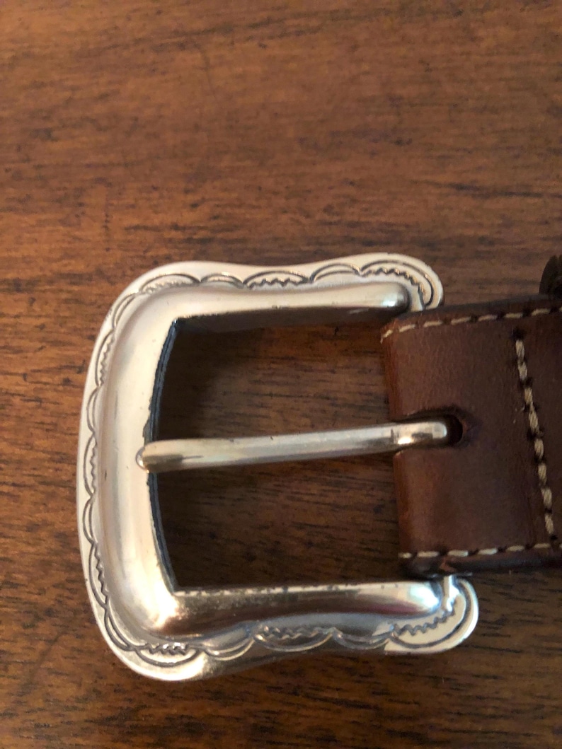 Puede incluir: Primer plano de una hebilla de cintur&oacute;n plateada con un borde festoneado decorativo. La hebilla est&aacute; sujeta a un cintur&oacute;n de cuero marr&oacute;n con costuras blancas. El dise&ntilde;o de la hebilla es de estilo western cl&aacute;sico.