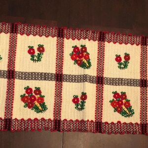 May include: A rectangular, woven table runner with a cream-colored background and a repeating pattern of red flowers. The runner has a red and black border and fringe on the ends. The flowers are embroidered in red and green.