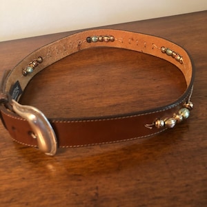 Puede incluir: Un cintur&oacute;n de cuero marr&oacute;n con hebilla plateada y detalles decorativos de cuentas. El cintur&oacute;n presenta una serie de peque&ntilde;as cuentas doradas y plateadas a lo largo del borde superior. La hebilla es de metal plateado. El cintur&oacute;n es para tallas de cintura entre 71 y 91 cm.