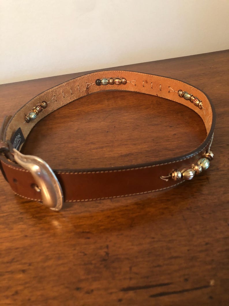 Puede incluir: Un cintur&oacute;n de cuero marr&oacute;n con hebilla plateada y detalles decorativos de cuentas. El cintur&oacute;n presenta una serie de peque&ntilde;as cuentas doradas y plateadas a lo largo del borde superior. La hebilla es de metal plateado. El cintur&oacute;n es para tallas de cintura entre 71 y 91 cm.