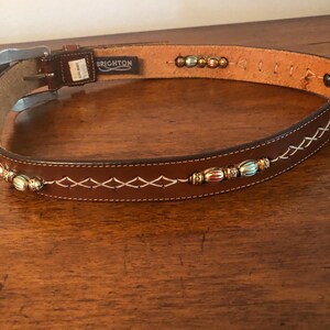 Puede incluir: Un cintur&oacute;n de cuero marr&oacute;n con costuras blancas y cuentas decorativas. El cintur&oacute;n tiene una hebilla plateada y la marca "BRIGHTON" es visible. El cintur&oacute;n est&aacute; sobre una superficie de madera.