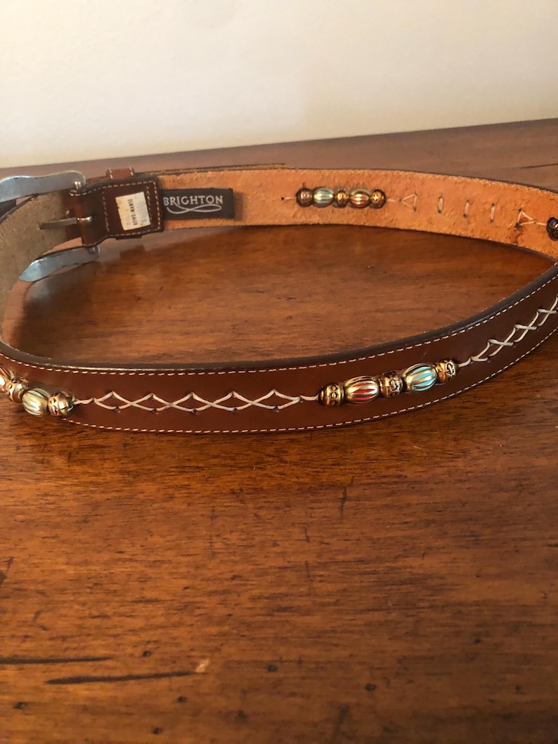 Puede incluir: Un cintur&oacute;n de cuero marr&oacute;n con costuras blancas y cuentas decorativas. El cintur&oacute;n tiene una hebilla plateada y la marca "BRIGHTON" es visible. El cintur&oacute;n est&aacute; sobre una superficie de madera.