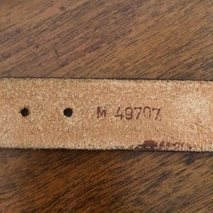 Puede incluir: Primer plano de un cintur&oacute;n de cuero marr&oacute;n claro con dos agujeros y el texto estampado "M 49707". El cintur&oacute;n est&aacute; sobre un fondo de madera.