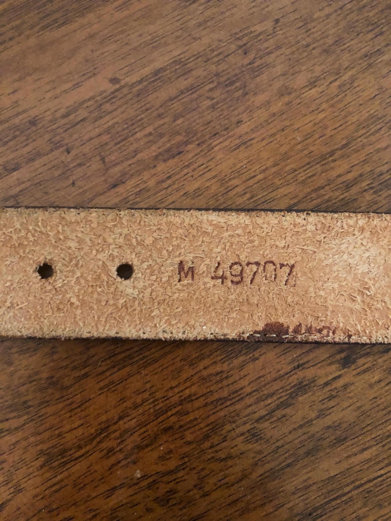 Puede incluir: Primer plano de un cintur&oacute;n de cuero marr&oacute;n claro con dos agujeros y el texto estampado "M 49707". El cintur&oacute;n est&aacute; sobre un fondo de madera.