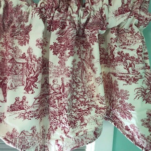 May include: Red and white toile fabric valance with a pattern of trees, houses, and people. The valance is gathered at the top and has a scalloped edge.