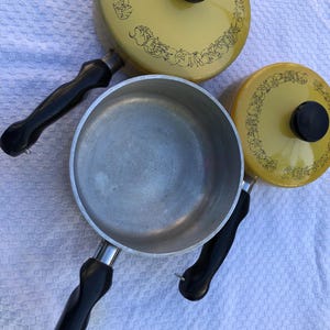 Op de afbeelding: Een set gele vintage pannen met zwarte handvatten en deksels. De deksels hebben een zwart decoratief patroon. De pannen zijn van metaal en staan op een witte ondergrond.