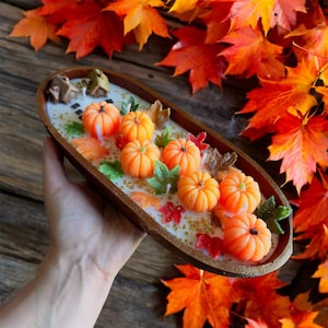 May include: A decorative wooden tray filled with orange pumpkin-shaped candles, green and red leaf accents, and a miniature house. The tray is surrounded by vibrant autumn leaves, creating a fall-themed display.