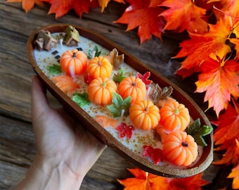 May include: A decorative wooden tray filled with orange pumpkin-shaped candles, green and red leaf accents, and a miniature house. The tray is surrounded by vibrant autumn leaves, creating a fall-themed display.