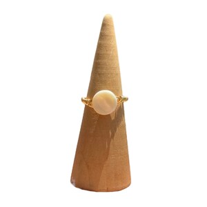 May include: A gold ring with a white round stone set in the center. The ring is displayed on a wooden ring holder.