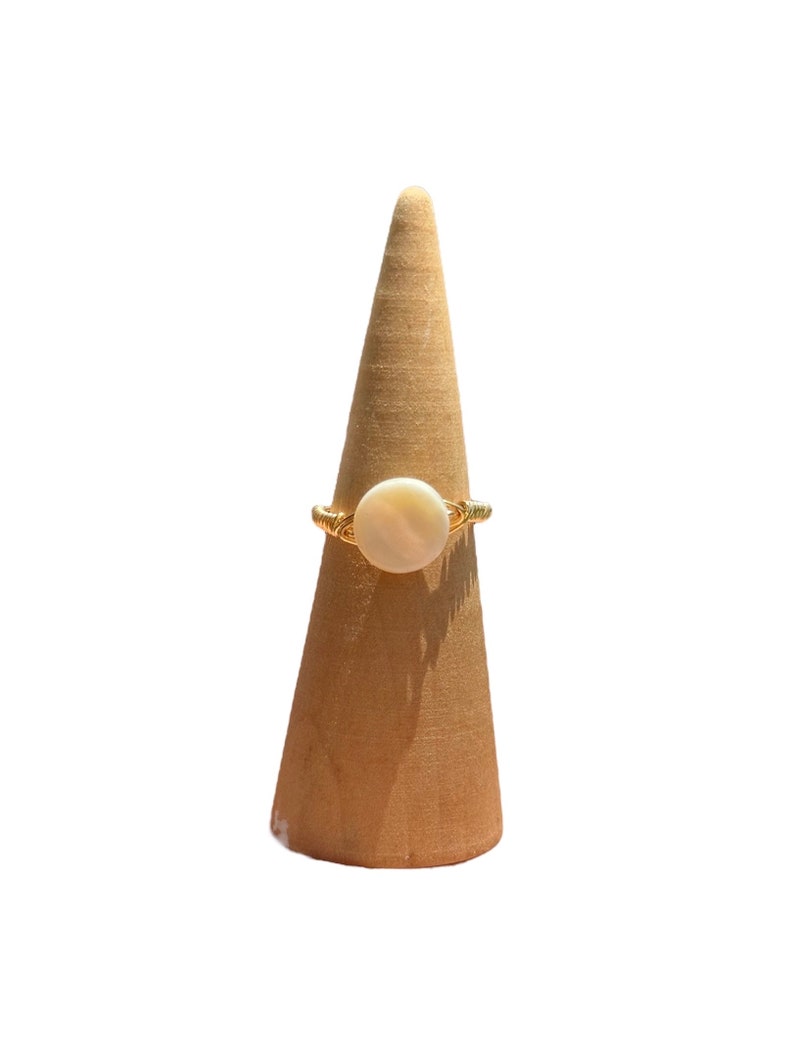 May include: A gold ring with a white round stone set in the center. The ring is displayed on a wooden ring holder.