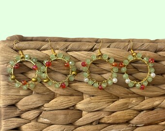 May include: Four pairs of gold hoop earrings, each adorned with green, red, and white beads. The earrings are displayed on a woven basket. The design suggests a handcrafted jewellery theme, with a delicate and intricate appearance.