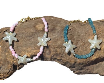 Puede incluir: Dos pulseras de cuentas con detalles dorados y abalorios de estrella de mar blanca. Una pulsera tiene cuentas rosas y la otra tiene cuentas azules.