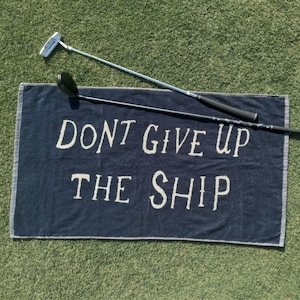 May include: A dark blue towel with the text "Don't Give Up The Ship" in white lettering. Two golf clubs are lying on top of the towel.
