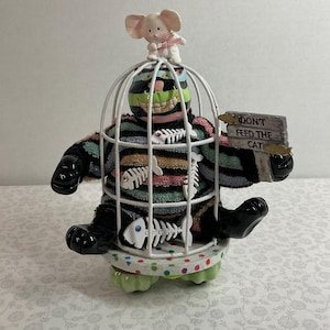 May include: A whimsical figurine of a creature in a cage, wearing a striped sweater and holding a sign that reads "DON'T FEED THE CAT." A small mouse figurine sits atop the cage. The base is decorated with polka dots and green accents.