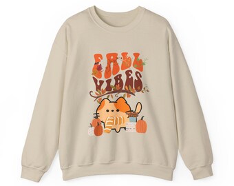 Op de afbeelding: Een beige sweatshirt met de woorden "Fall Vibes" in oranje, met een cartoonkat en pompoenen. De kat is oranje en wit, met een gestreepte sjaal. Pompoenen en herfstbladeren zijn ook afgebeeld.