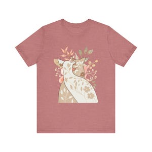 Peut inclure: Un t-shirt rose poudré à manches courtes avec un motif floral représentant deux chats stylisés. Les chats sont de couleur crème et beige, avec des accents floraux et de feuilles pêche, verts et bruns. Le motif est centré sur le t-shirt.