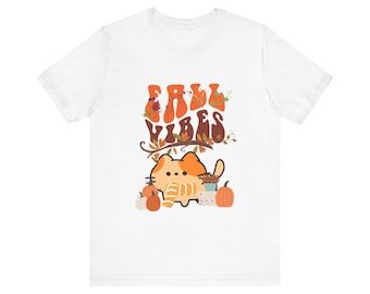 Könnte beinhalten: Weißes T-Shirt mit einer süßen orangefarbenen Katze, die einen Schal trägt und von Kürbissen und Herbstblättern umgeben ist. Der Text "Fall Vibes" ist in einer Retro-Schrift über der Katze geschrieben.