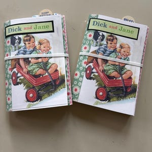 May include: Two identical small booklets featuring a vintage illustration of two children in a red wagon with a dog. The cover displays the text "Dick and Jane" in a green box. Each booklet is secured with a white elastic band and has a small tab.