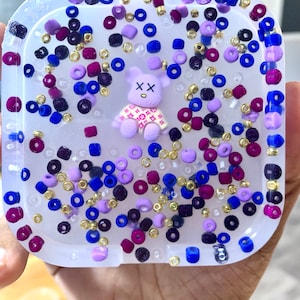 May include: A clear resin tray with a purple teddy bear wearing a pink and white patterned shirt. The tray is filled with colorful beads in shades of purple, blue, gold, and white.