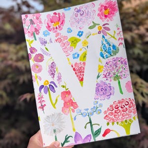 Peut inclure: Une feuille de papier rectangulaire présentant une grande lettre V blanche en espace négatif, entourée d'une illustration aquarelle vibrante de diverses fleurs roses, violettes, jaunes et bleues avec des tiges et feuilles vertes.